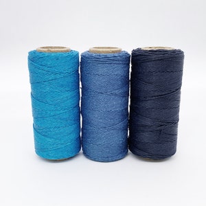 May include: Three spools of thread in shades of blue. The spools are wound with a thick, textured thread. The thread is a light blue, a medium blue, and a dark blue.