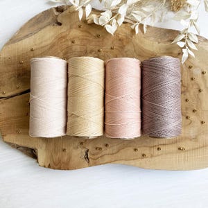 Könnte beinhalten: Vier Spulen Makramee-Garn in neutralen Farbtönen wie Beige, Hellbeige und Braun, auf einem Holzbrett angeordnet. Die eng gewickelten Garne zeigen ihre Textur. Getrocknete Blumenelemente und kleine goldene Perlen ergänzen die natürliche Ästhetik.