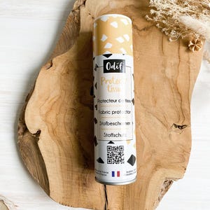 Peut inclure: Un aérosol protecteur de tissu cylindrique avec un motif géométrique jaune et blanc sur le dessus. L'étiquette comprend la marque "Odif" et le texte "Protege tissu" et "Fabric protector". Le flacon est posé sur une surface en bois.