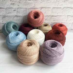 Könnte beinhalten: Zehn Knäuel Garn in verschiedenen Farben, darunter Beige, Blau, Braun, Creme, Grau, Lavendel und Rot. Die Knäuel Garn sind in einem Kreis auf einer weißen Oberfläche angeordnet.