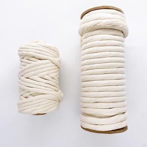 May include: Two spools of cream-coloured cotton rope. One spool is shorter and wider, the other is taller and thinner. Both have a wooden base. The rope is tightly wound, creating a textured surface. Suitable for macrame, crafting, or decorative use.