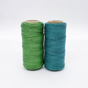 May include: Two spools of thread, one green and one teal, are side by side on a white surface.