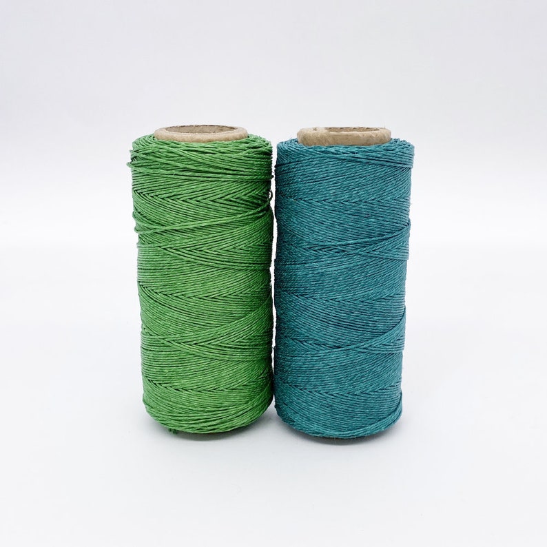 May include: Two spools of thread, one green and one teal, are side by side on a white surface.