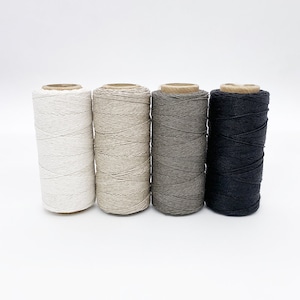 May include: Four spools of natural fiber twine in white, beige, gray, and black. The spools are arranged in a row from left to right.