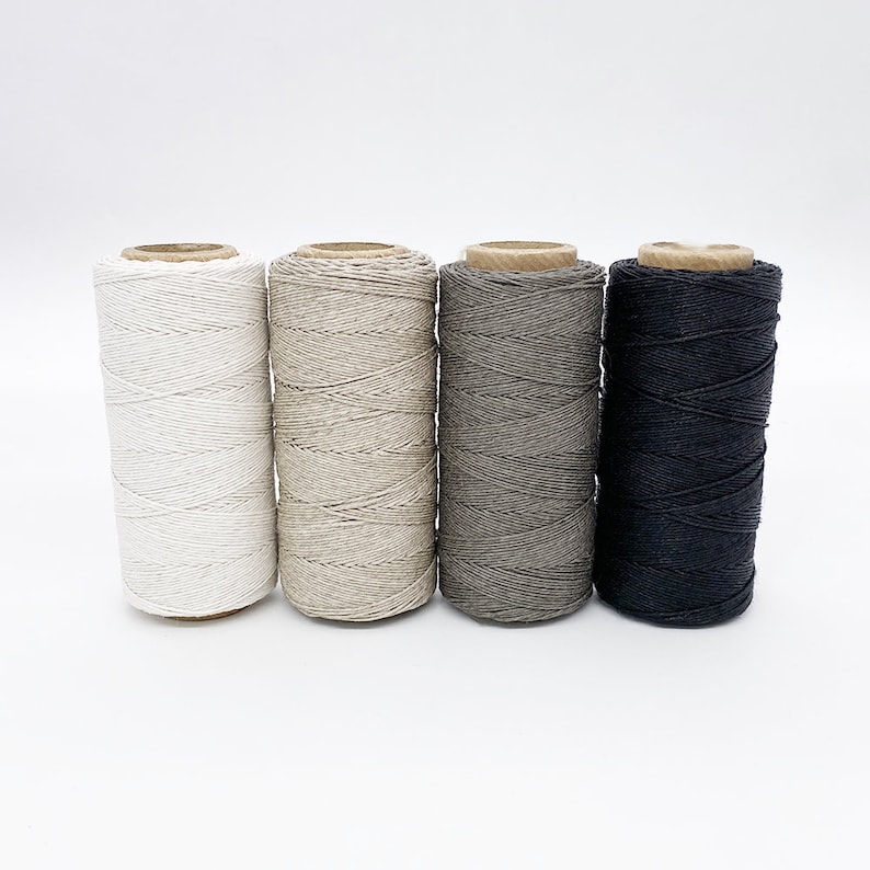 May include: Four spools of natural fiber twine in white, beige, gray, and black. The spools are arranged in a row from left to right.