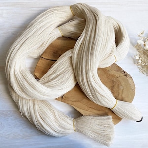 Puede incluir: Hilo de fibra natural blanquecino, agrupado y atado con bandas amarillas. El hilo está dispuesto en un patrón en bucle sobre una superficie de madera, mostrando su textura y color. La imagen resalta el aspecto crudo y sin procesar del material.