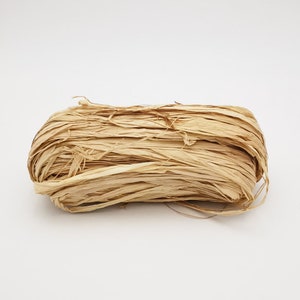 May include: A close-up of a bundle of natural, beige paper raffia. The raffia is loosely packed and has a slightly textured appearance.