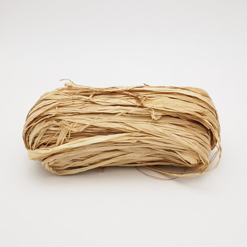May include: A close-up of a bundle of natural, beige paper raffia. The raffia is loosely packed and has a slightly textured appearance.