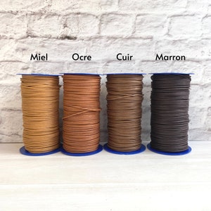 May include: Four spools of leather cord in different colours: miel, ocre, cuir, and marron. The spools are arranged in a row on a white wooden surface.