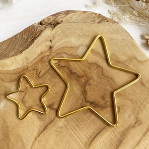 May include: Two gold metal star shapes of different sizes on a wooden surface. The larger star is about 25 cm wide and the smaller star is about 12.5 cm wide.