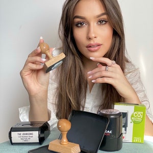 May include: A woman holds a wooden stamp with a black ink pad. There are three other ink pads and stamps on a table, including one with the text "THE JOHNSONS 1234 Northern Street New York, NY +10022" and another with the text "SEAL DOR".