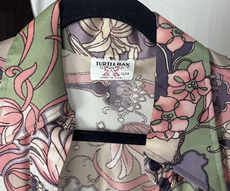 May include: A vintage button-down shirt with a floral pattern in shades of pink, green, and purple. The shirt features a collar and a label that reads "TURTLE BAX" and "13/14". Made in the USA.