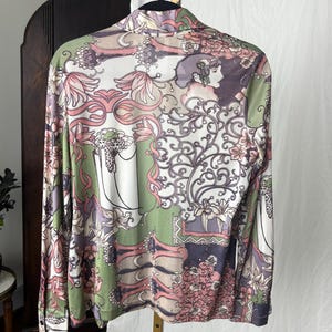 May include: A long-sleeved button-down shirt with a colorful floral and art nouveau design. The shirt features a mix of pink, green, white, and purple hues, with a detailed pattern of flowers, vines, and a woman's profile.