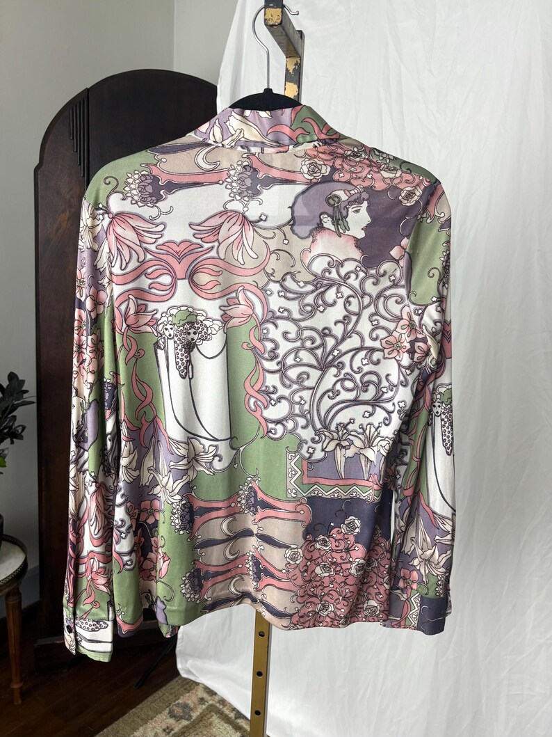 May include: A long-sleeved button-down shirt with a colorful floral and art nouveau design. The shirt features a mix of pink, green, white, and purple hues, with a detailed pattern of flowers, vines, and a woman's profile.