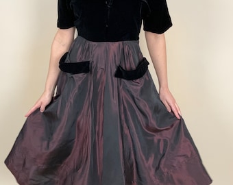 May include: A black velvet bodice with short sleeves and a button closure, paired with a full, iridescent taffeta skirt. The skirt features two large pockets with black velvet trim.
