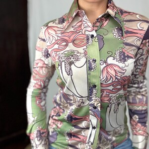 May include: A button-down shirt with a floral pattern in shades of pink, green, and purple. The long-sleeved shirt has a pointed collar and is made of a silky material. The shirt is paired with blue jeans.