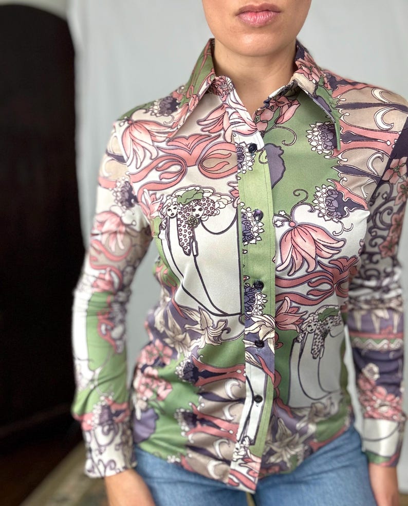 May include: A button-down shirt with a floral pattern in shades of pink, green, and purple. The long-sleeved shirt has a pointed collar and is made of a silky material. The shirt is paired with blue jeans.