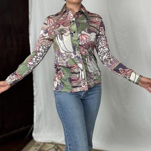 May include: A button-down shirt with a floral pattern in shades of green, pink, and white. The long-sleeved shirt has a pointed collar and is paired with blue jeans. The shirt's design features stylized floral and leaf motifs.