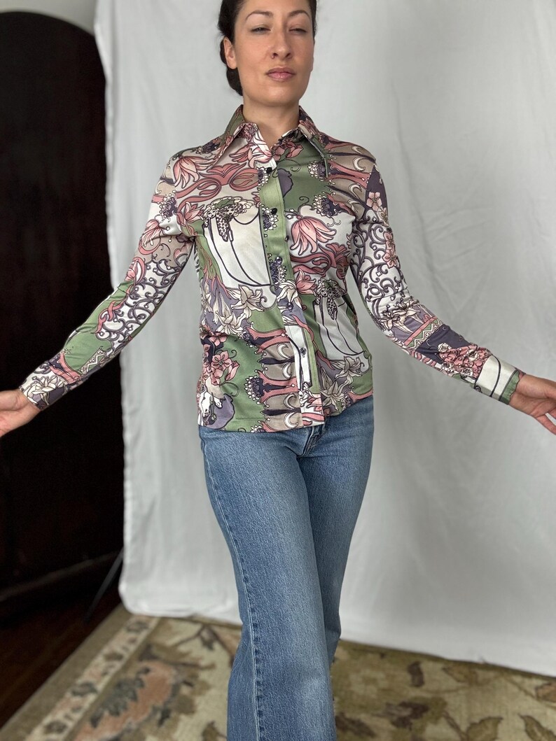 May include: A button-down shirt with a floral pattern in shades of green, pink, and white. The long-sleeved shirt has a pointed collar and is paired with blue jeans. The shirt's design features stylized floral and leaf motifs.