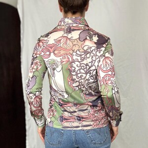 May include: A long-sleeved button-down shirt with a colorful floral and figure pattern in shades of pink, green, purple, and beige. The shirt has a collared neckline and buttoned cuffs. The fabric appears to be a silky material.