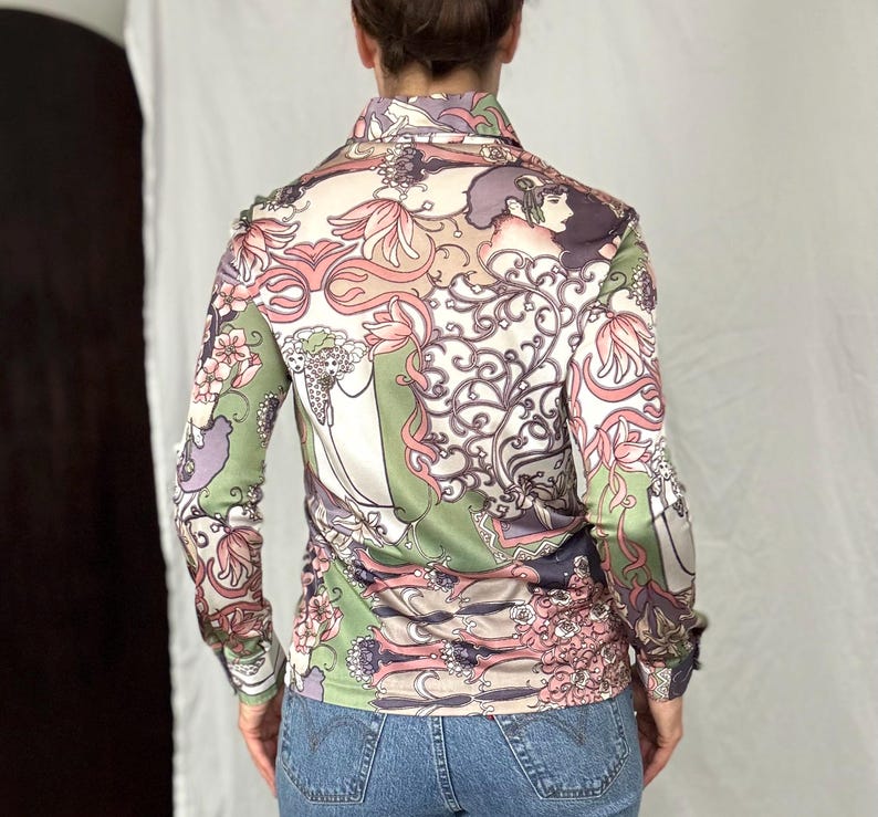 May include: A long-sleeved button-down shirt with a colorful floral and figure pattern in shades of pink, green, purple, and beige. The shirt has a collared neckline and buttoned cuffs. The fabric appears to be a silky material.