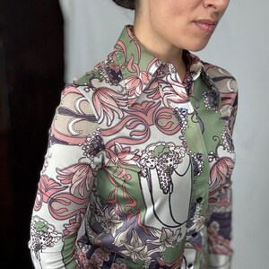 May include: A button-down shirt with a colorful floral pattern in shades of pink, green, and white. The long-sleeved shirt has a collared neckline and a silky texture. The design features stylized flowers and swirling shapes.