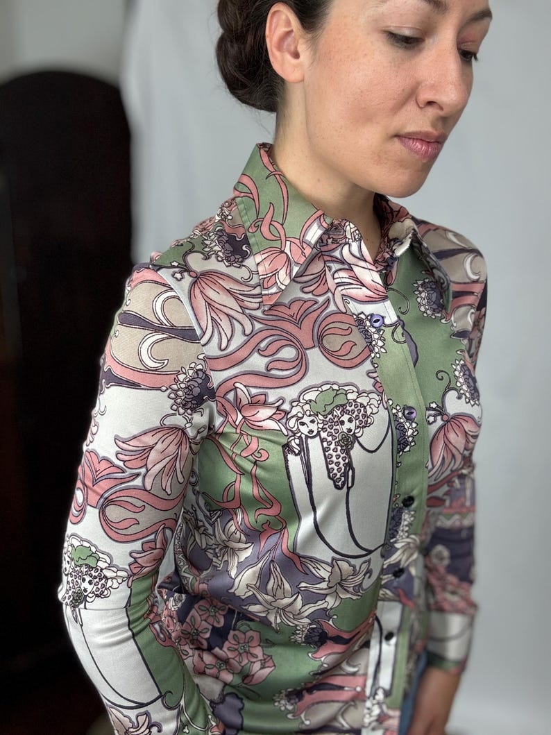 May include: A button-down shirt with a colorful floral pattern in shades of pink, green, and white. The long-sleeved shirt has a collared neckline and a silky texture. The design features stylized flowers and swirling shapes.