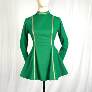 May include: A green long-sleeved dress with a high neckline and a flared skirt. Gold trim accents the neckline and runs down the front of the dress. The dress is displayed on a mannequin.