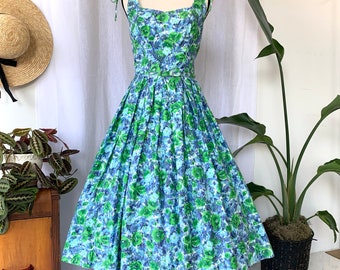 May include: A vintage-style floral dress in shades of blue and green. The dress features a fitted bodice with tie straps and a full, pleated skirt. A matching belt cinches the waist. The dress is displayed on a mannequin, showcasing its classic silhouette.