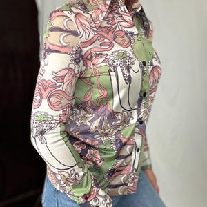May include: A button-down shirt with a floral pattern in shades of pink, green, and white. The long-sleeved shirt has a collared neckline and black buttons. The floral design includes stylized flowers and swirling lines. The shirt is paired with blue jeans.