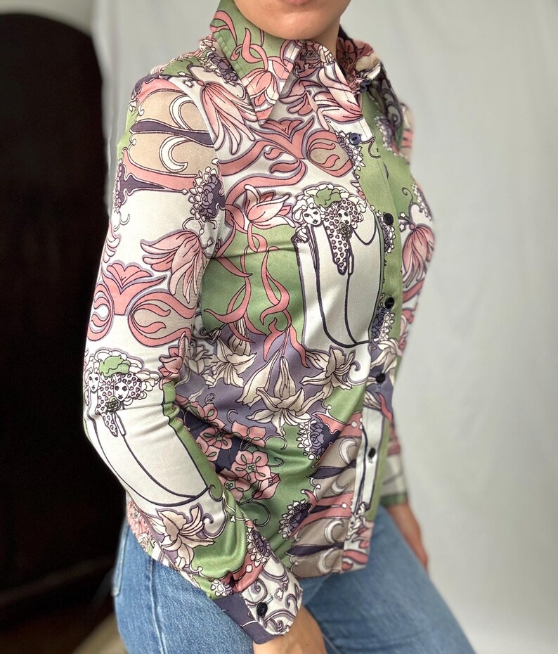 May include: A button-down shirt with a floral pattern in shades of pink, green, and white. The long-sleeved shirt has a collared neckline and black buttons. The floral design includes stylized flowers and swirling lines. The shirt is paired with blue jeans.