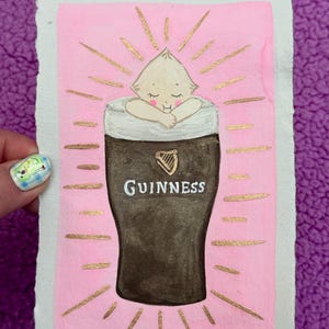 Puede incluir: Una pintura de acuarela caprichosa sobre papel texturizado. La obra presenta una figura parecida a un beb&eacute; acurrucada en un vaso de pinta Guinness, con la palabra "Guinness" visible. El fondo es rosa con l&iacute;neas doradas radiantes.