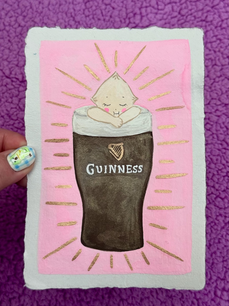 Puede incluir: Una pintura de acuarela caprichosa sobre papel texturizado. La obra presenta una figura parecida a un beb&eacute; acurrucada en un vaso de pinta Guinness, con la palabra "Guinness" visible. El fondo es rosa con l&iacute;neas doradas radiantes.