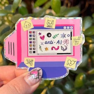 Op de afbeelding: Een roze en blauwe computersticker met de tekst "anti-AI" op het scherm. De sticker heeft verschillende plaknotities met tekeningen van een kat, een gezicht en een hart. Andere afbeeldingen zijn een ster, een cursor en het cijfer 8.