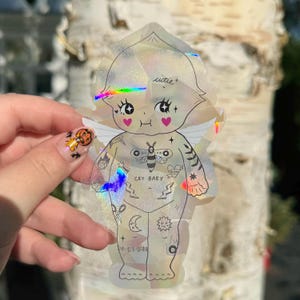Puede incluir: Pegatina holográfica de una figura querubínica con alas, tatuajes y las palabras "cutie" y "cry baby". La pegatina tiene un brillo de arcoíris y presenta una polilla estilizada y detalles de corazón. El diseño se sostiene contra un fondo borroso.