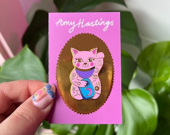 May include: A pink and gold enamel pin featuring a cartoon cat with a blue and purple collar and tattoos. The cat is holding a blue and gold crescent moon. The pin is on a pink card with the text "Amy Hastings" in black.