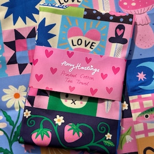 Puede incluir: Un paño de cocina de algodón plegado con un vibrante diseño de patchwork de corazones, setas, fresas y estrellas. Una banda de papel rosa envuelve el centro, impresa con el texto Amy Hastings Printed Cotton Tea Towel y corazones pequeños.