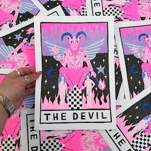 Könnte beinhalten: Eine rosa und schwarze Tarotkarten-Illustration des Teufels. Der Teufel ist ein rosa humanoid mit Hörnern, Flügeln und einem Schwanz. Der Teufel sitzt auf einer karierten Plattform mit zwei Figuren, die auf beiden Seiten stehen. Die Karte ist von Flammen umgeben.