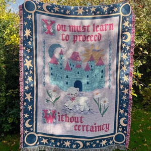 Könnte beinhalten: Eine gewebte Wurfadecke mit einem skurrilen Design. Die Decke hat einen hellvioletten Hintergrund mit einem Schloss, einem Lamm und floralen Illustrationen. Der Text lautet: "You must learn to proceed without certainty." Der Rand ist marineblau mit Sternen und Monden.