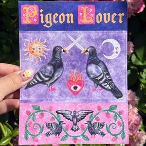 May include: A rectangular art print featuring the text Pigeon Lover in yellow letters on a blue background. The illustration shows two grey pigeons facing each other with crossed swords, a sun, a moon, and a flaming heart with an eye.