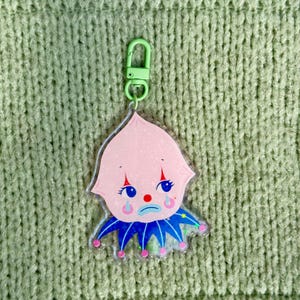 May include: A pink and blue cartoon clown keyring with a sad expression. The keyring is made of clear acrylic and has a green metal clasp.