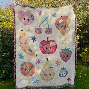 May include: A cream-coloured throw blanket featuring whimsical illustrations of fruits with faces, including an apple, banana, and strawberry. The blanket has a fringed edge and is decorated with star shapes in various colours.