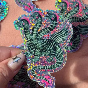 Könnte beinhalten: Ein holografischer Aufkleber, der ein dreiköpfiges grünes Wesen mit Flügeln und einem langen Schwanz zeigt. Das Wesen hat ein strukturiertes Muster auf dem Körper, rosa Akzente an Wangen und Schwanz und ist von einem schimmernden Glitzereffekt umgeben.