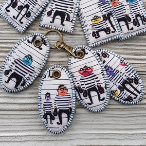 Könnte beinhalten: Satz von sieben bunten Schlüsselanhängern mit einer Comicfigur in einem gestreiften Gefängnisuniform mit einer Geldbörse. Die Figur steht vor einem weißen Hintergrund mit schwarzen Linien und Zahlen, die an eine Polizeiaufstellung erinnern.