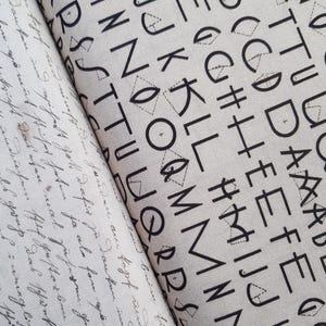 Puede incluir: Dos muestras de tela con diferentes patrones. Una presenta letras negras y formas geométricas sobre un fondo claro. La otra tiene escritura manuscrita en tinta negra sobre un fondo claro. Ambas telas están dobladas.