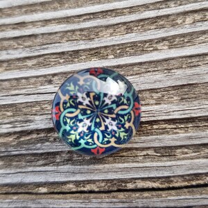 Op de afbeelding: Een ronde glazen cabochon met een kleurrijk, ingewikkeld ontwerp. Het ontwerp heeft een centraal motief met draaiende patronen in tinten blauw, groen, geel en rood.