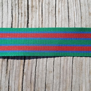 Op de afbeelding: Een close-up van een groene geweven band met twee rode en twee blauwe strepen. De band ligt op een verweerd houten oppervlak. De band wordt waarschijnlijk gebruikt voor knutselen of als decoratief element.