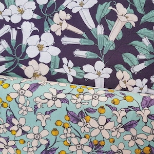 Könnte beinhalten: Zwei Stoffmuster mit Blumenmustern. Das obere Muster ist ein dunkelvioletter Stoff mit weißen Blüten und grünen Blättern. Das untere Muster ist ein hellblauer Stoff mit weißen Blüten, violetten Blättern und gelben Beeren.