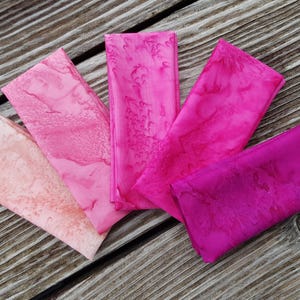 Op de afbeelding: Vijf gevouwen stoffen servetten in roze tinten, van licht perzik tot diep magenta, gerangschikt op een verweerd houten oppervlak. De servetten hebben een gemarmerd of aquarelachtig patroon.