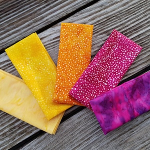 Peut inclure: Cinq carrés de tissu colorés dans les tons de jaune, orange, rose et violet sont disposés sur une surface en bois vieilli. Les tissus présentent divers motifs, dont des pois et des effets tie-dye, créant un affichage vibrant.